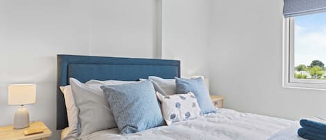 Un lit bien fait, avec une literie bleue et blanche, une table de chevet avec une lampe et une fenêtre donnant sur un paysage verdoyant.