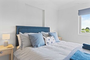 A neatly made bed with blue and white bedding, a bedside table with a lamp, and a window with a view of greenery. - Stunning Beachfront Bungalow  Ocean Views & BBQ (Pevensey Bay)
