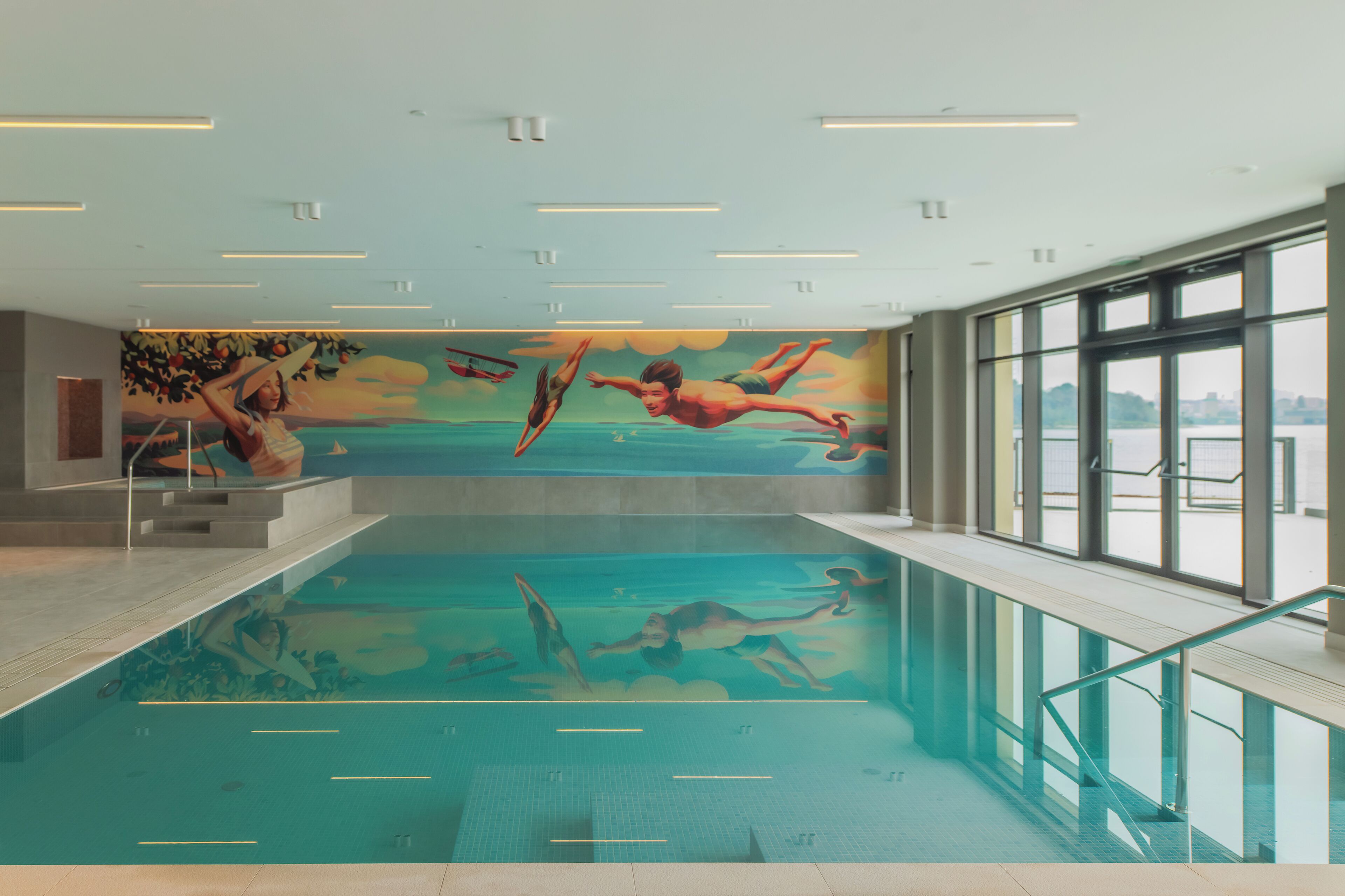 An indoor swimming pool with a mural of swimmers, large windows, and a clear view of the outside.
