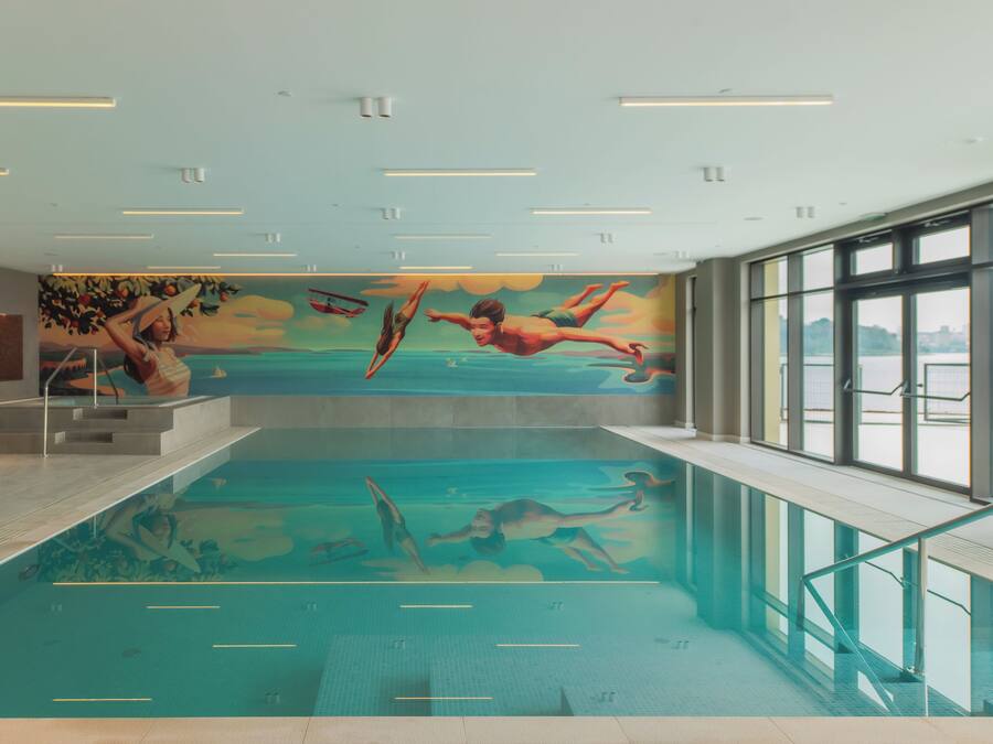 Una piscina interior con un mural de nadadores, ventanas amplias y vistas despejadas al exterior.