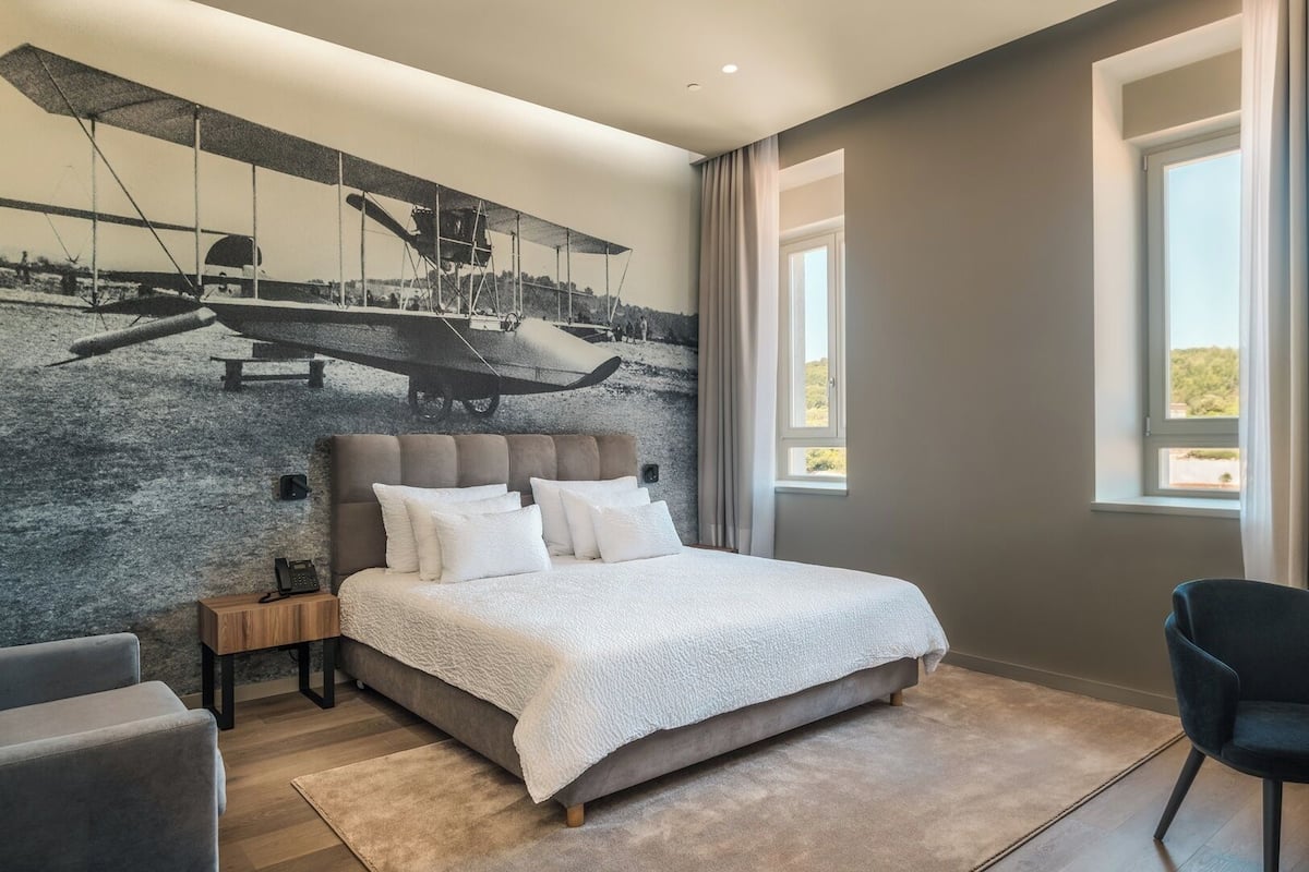 Une chambre avec un grand lit, une table de chevet, une chaise, une fenêtre et une fresque représentant un avion sur le mur.