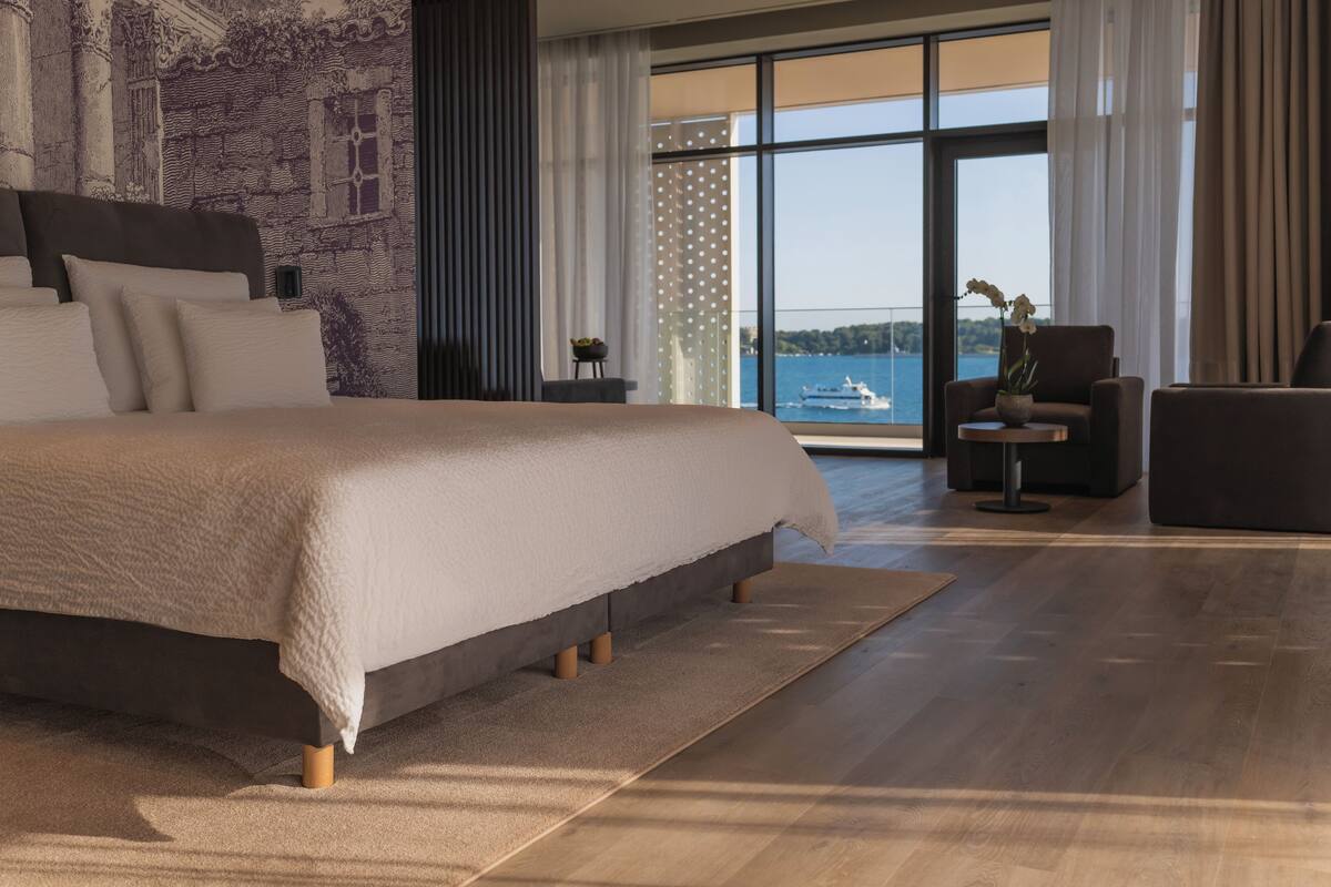 Un grand lit avec une tête de lit, une petite table avec une plante et une vue sur la mer par de grandes fenêtres.