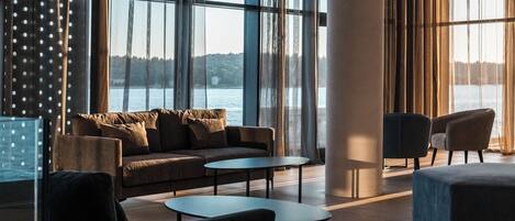 A modern lounge area with a sofa, two round tables, and chairs, large windows with curtains, and a view of a body of water.