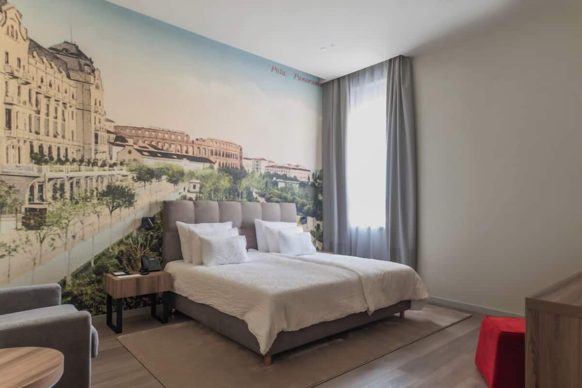 Une chambre d’hôtel avec un grand lit, une table de chevet, un fauteuil rouge et une fresque représentant un paysage urbain sur le mur.