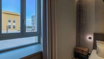 Una habitación de hotel con un ventanal grande, una mesita de noche, una cama y una lámpara fijada a la pared.