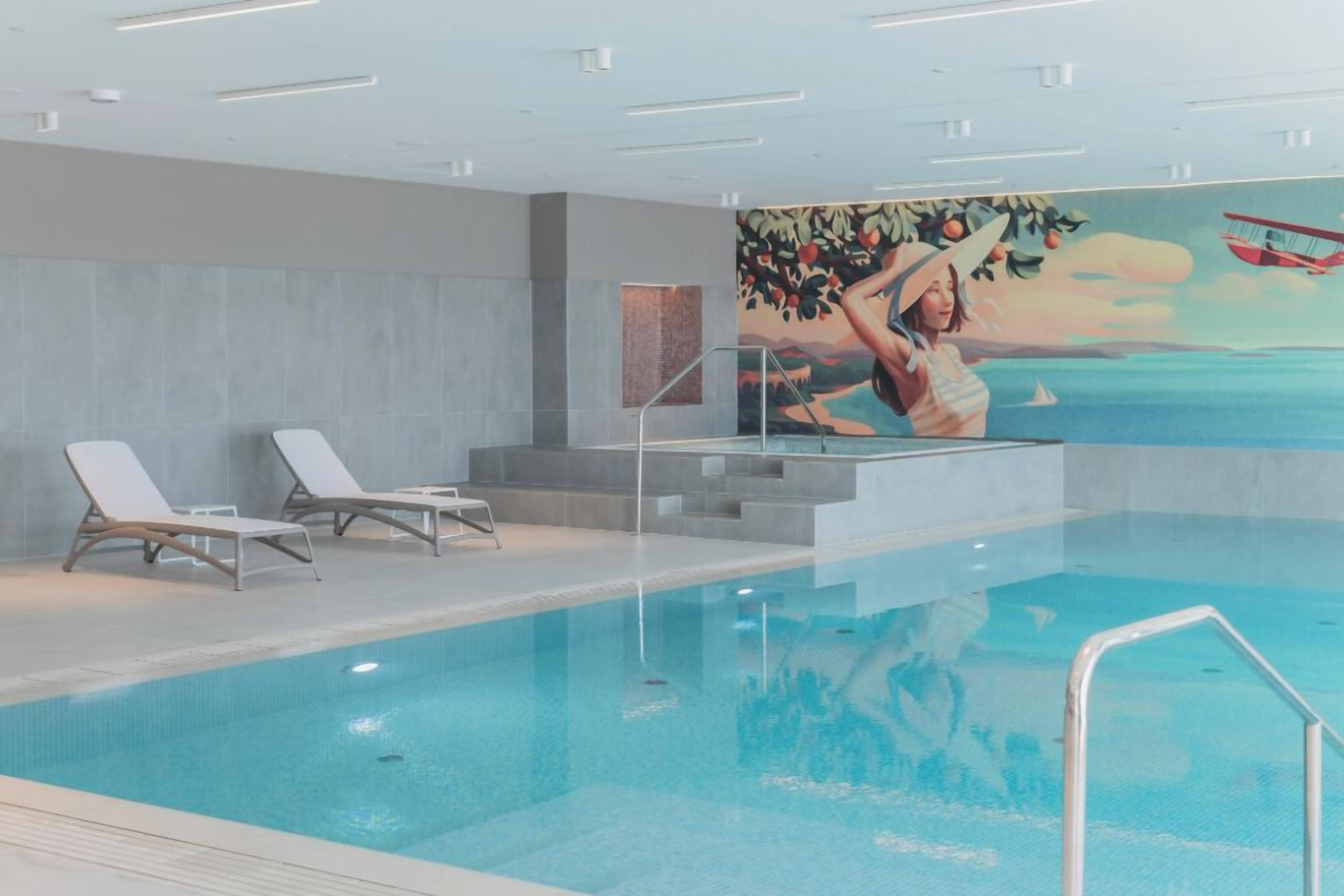 A modern indoor swimming pool with clear water, white lounge chairs, and a mural depicting a woman in a hat.