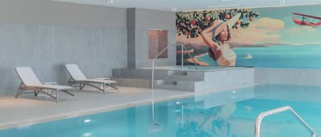 Piscina interior moderna con agua cristalina, sillas de descanso blancas y un mural que muestra a una mujer con sombrero.