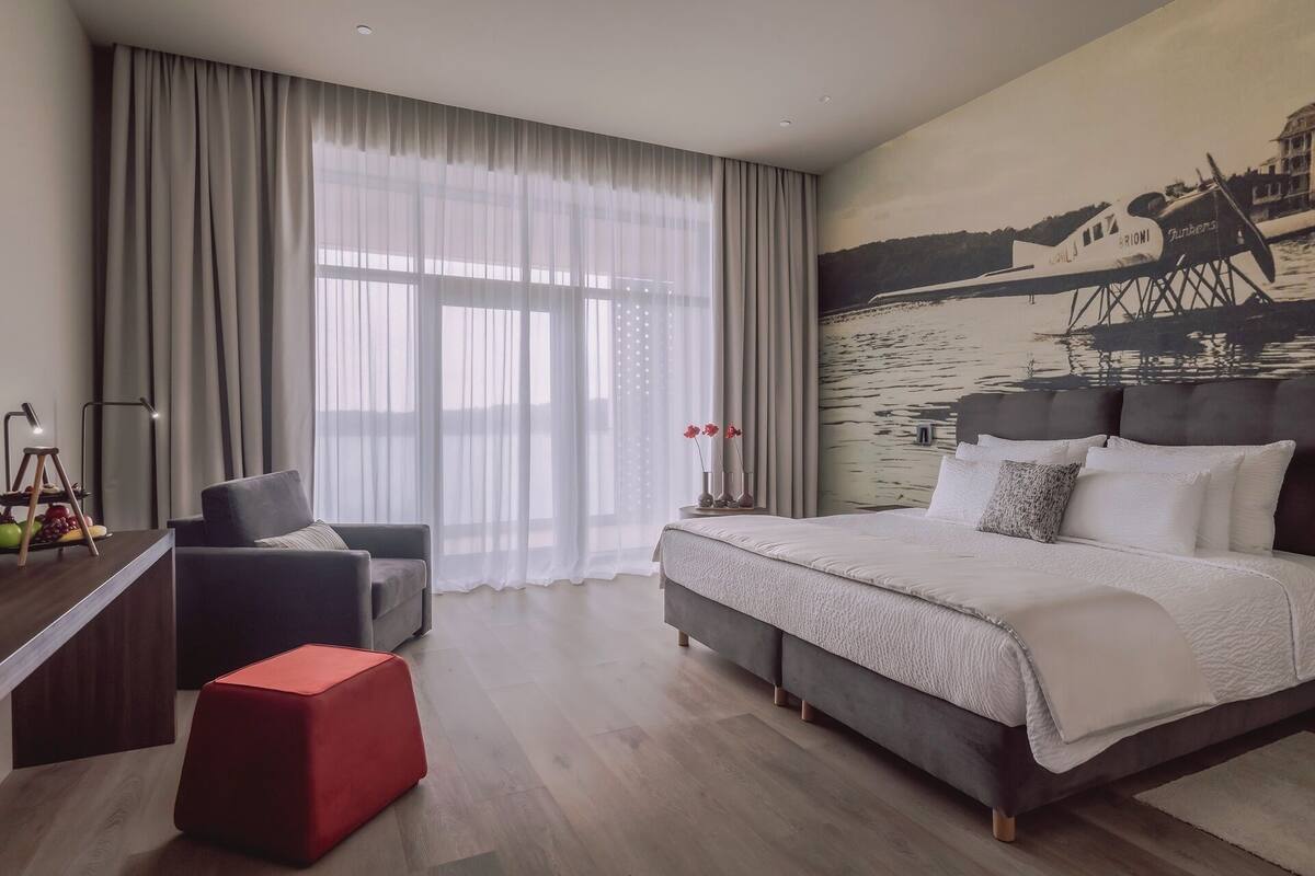 Une chambre d’hôtel moderne avec un grand lit, un pouf rouge, un canapé gris et un tableau représentant un bateau accroché au mur.