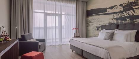 Una habitación de hotel moderna con una cama grande, un reposapiés rojo, un sofá gris y un cuadro de un barco en la pared.