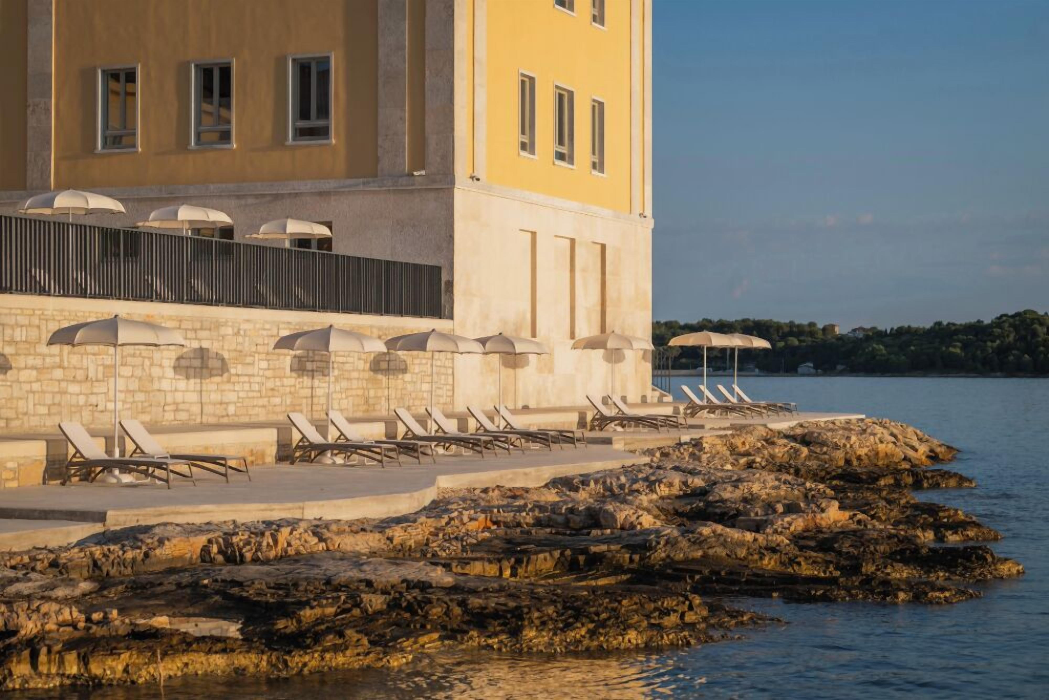 A coastal hotel with a stone facade, outdoor seating with umbrellas, and a rocky shoreline.