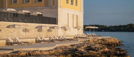 A coastal hotel with a stone facade, outdoor seating with umbrellas, and a rocky shoreline.