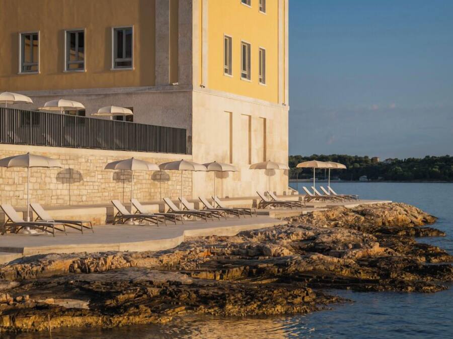 A coastal hotel with a stone facade, outdoor seating with umbrellas, and a rocky shoreline.