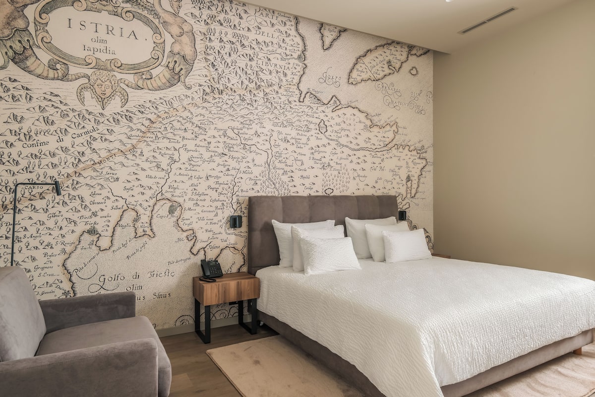 Une chambre avec une grande fresque représentant une carte géographique au mur, un lit avec une literie blanche, une table de chevet et une chaise.