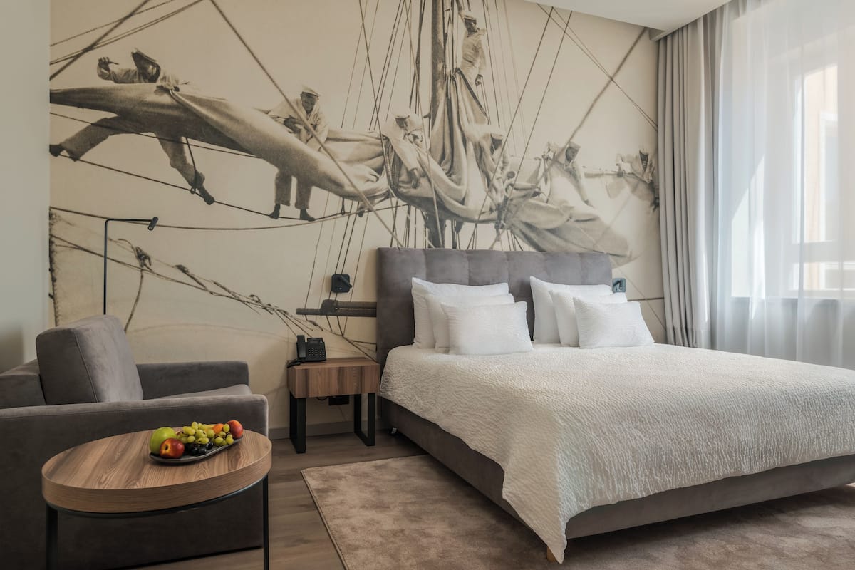Une chambre d’hôtel moderne avec un grand lit, un canapé gris, une petite table ronde avec des fruits et une fresque murale représentant un équipage de voilier.