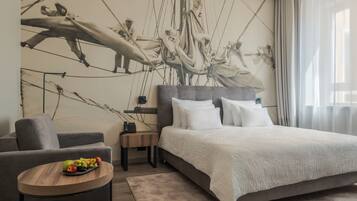 A modern hotel room with a large bed, a grey sofa, a small round table with fruit, and a mural of a sailing crew on the wall.