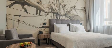 A modern hotel room with a large bed, a grey sofa, a small round table with fruit, and a mural of a sailing crew on the wall.
