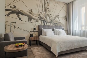 Une chambre d’hôtel moderne avec un grand lit, un canapé gris, une petite table ronde avec des fruits et une fresque murale représentant un équipage de voilier.