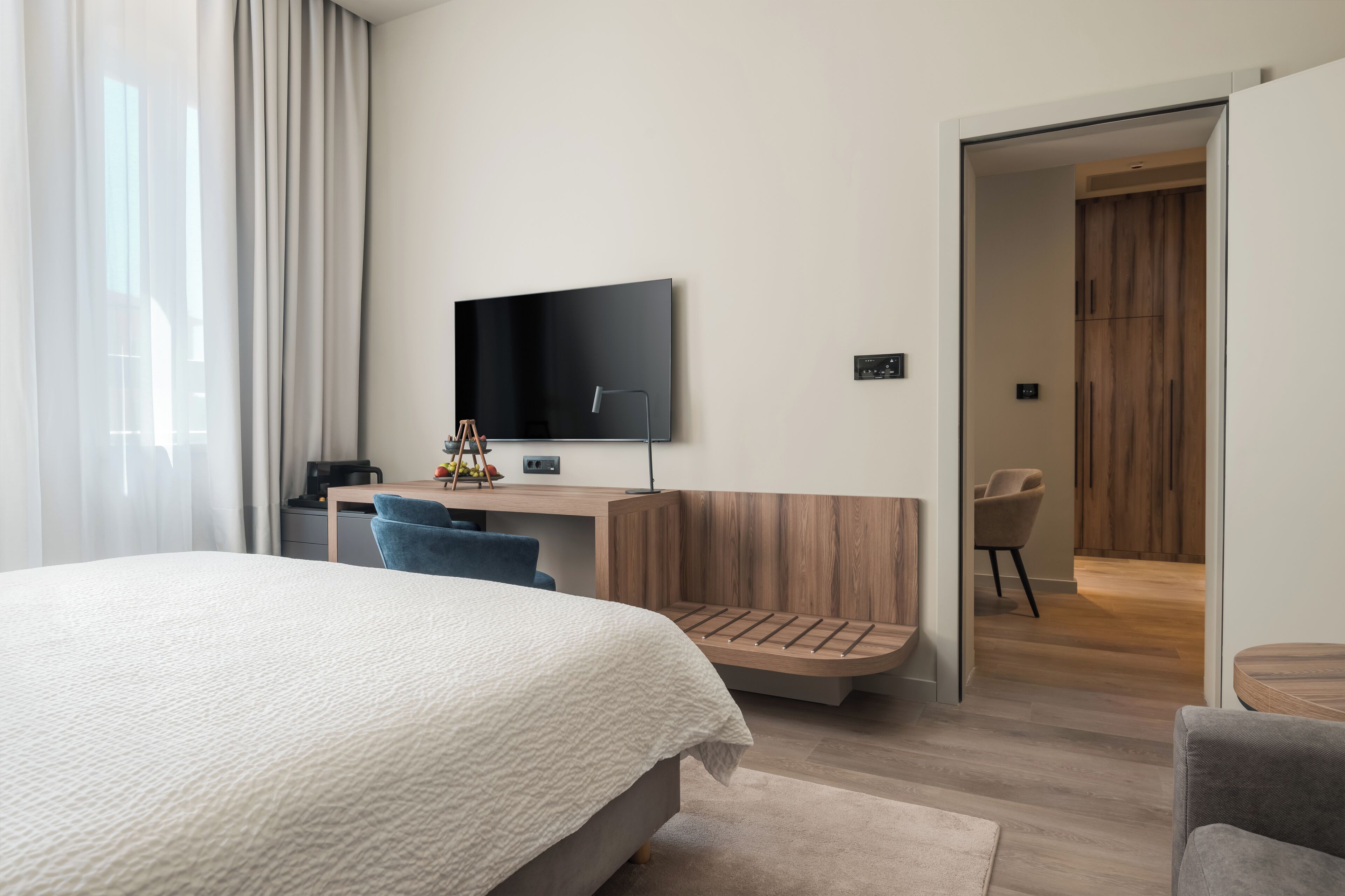 A modern hotel room with a large bed, a desk with a flat-screen TV, a wooden bench, and a chair.