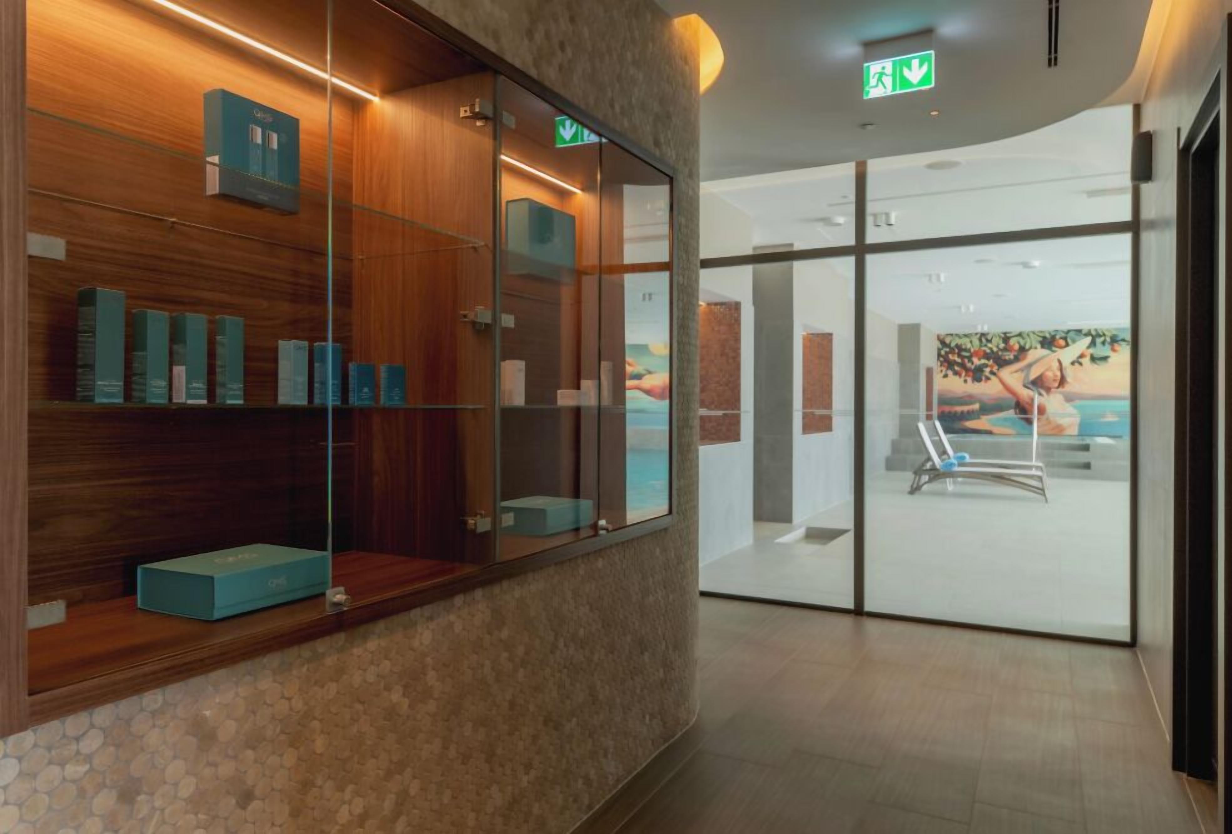 Een hotel receptie met een houten balie, een glazen scheidingswand en uitzicht op een zwembad met een ligstoel.