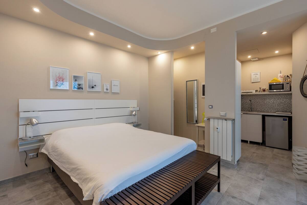 Un dormitorio moderno con una cama grande, mesitas de noche, un banco y una kitchenette al fondo.