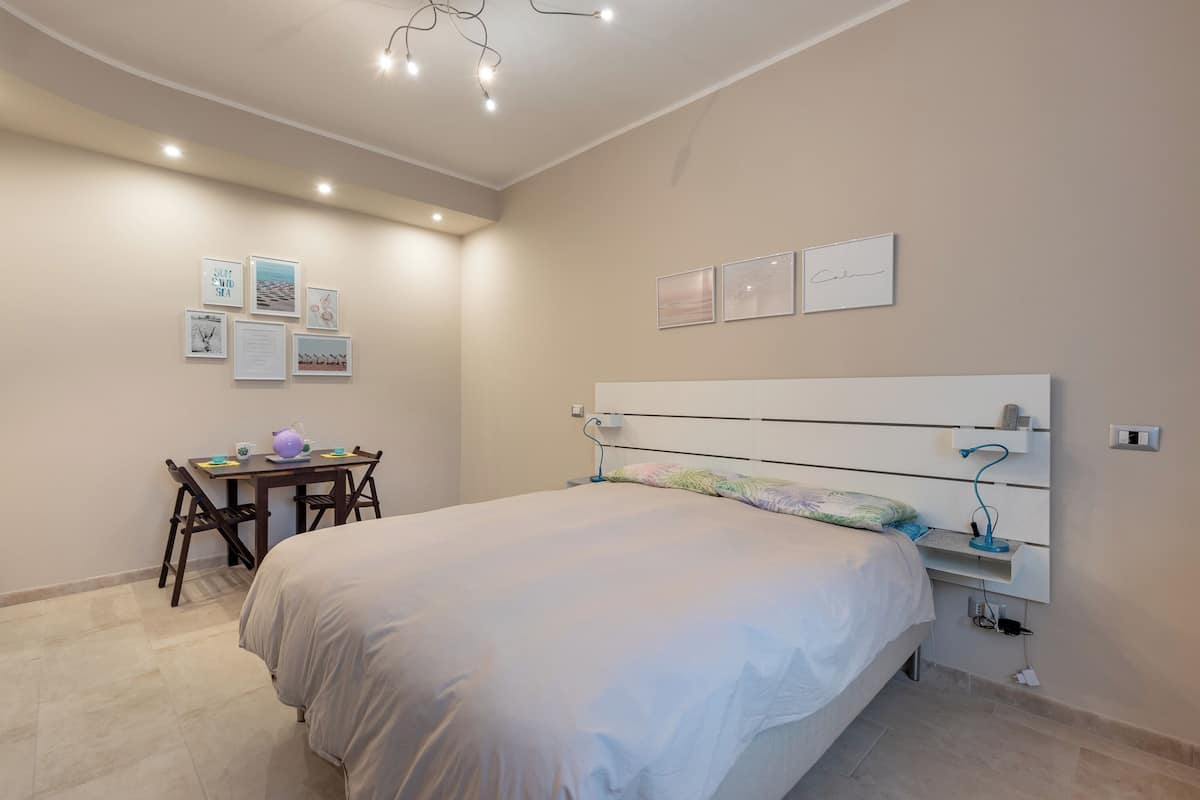 Un dormitorio con cama, escritorio y silla. Hay luces fijadas a la pared y cuadros enmarcados.