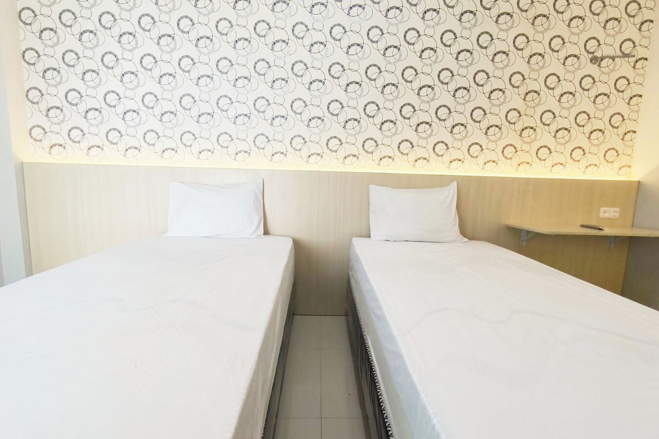 A hotel room with two single beds, a wooden headboard, a patterned wall, and a bedside table with a lamp.