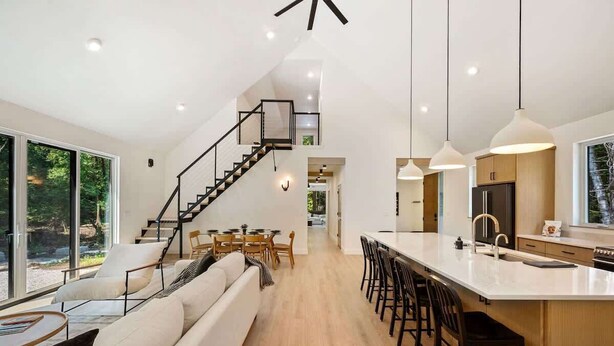 A modern kitchen with a central island, a dining area with a staircase leading to an upper level, and a large window providing a view of the outdoors. - Modern Luxury Retreat · Game Loft · Near Bay (Egg Harbor)