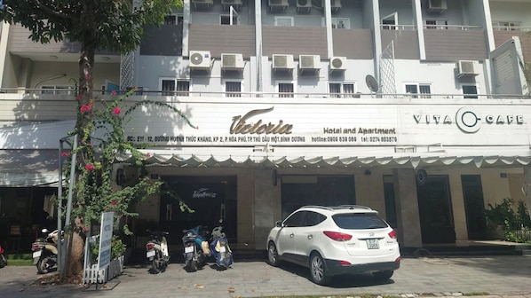 Un hotel llamado Victoria con un letrero que indica que ofrece servicios de hotel y apartamentos. Hay coches y motocicletas estacionados al frente, y un árbol con flores rosadas a la izquierda.