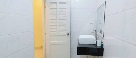 A small, narrow bathroom with white tiled walls and floor, a white toilet, a black sink with a mirror above it, and a door leading to another room.