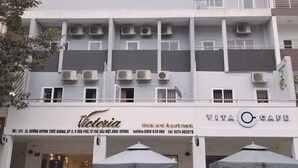 Un edificio con un letrero que indica 'Victoria Hotel and Apartment' y 'Vita Cafe'. Se observan unidades de aire acondicionado en el exterior, y dos sombrillas están dispuestas frente al café.