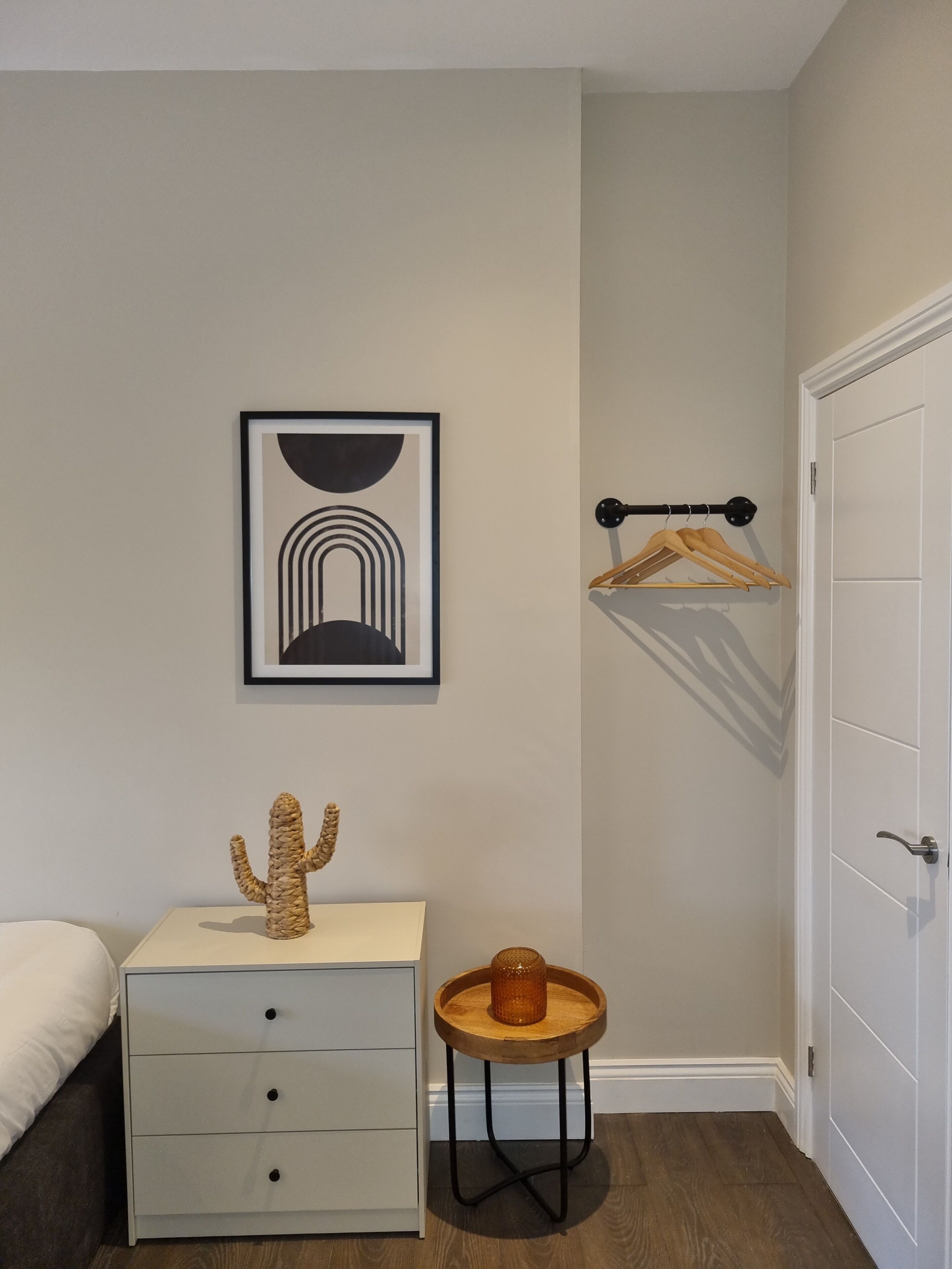 A bedroom with a bed, a dresser, a framed artwork, a small wooden table with a vase, and a coat rack.