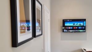 A hallway with framed artwork on the wall, a television mounted above a door, and a chair in the corner. - The Ivy Apartments (Bournemouth)