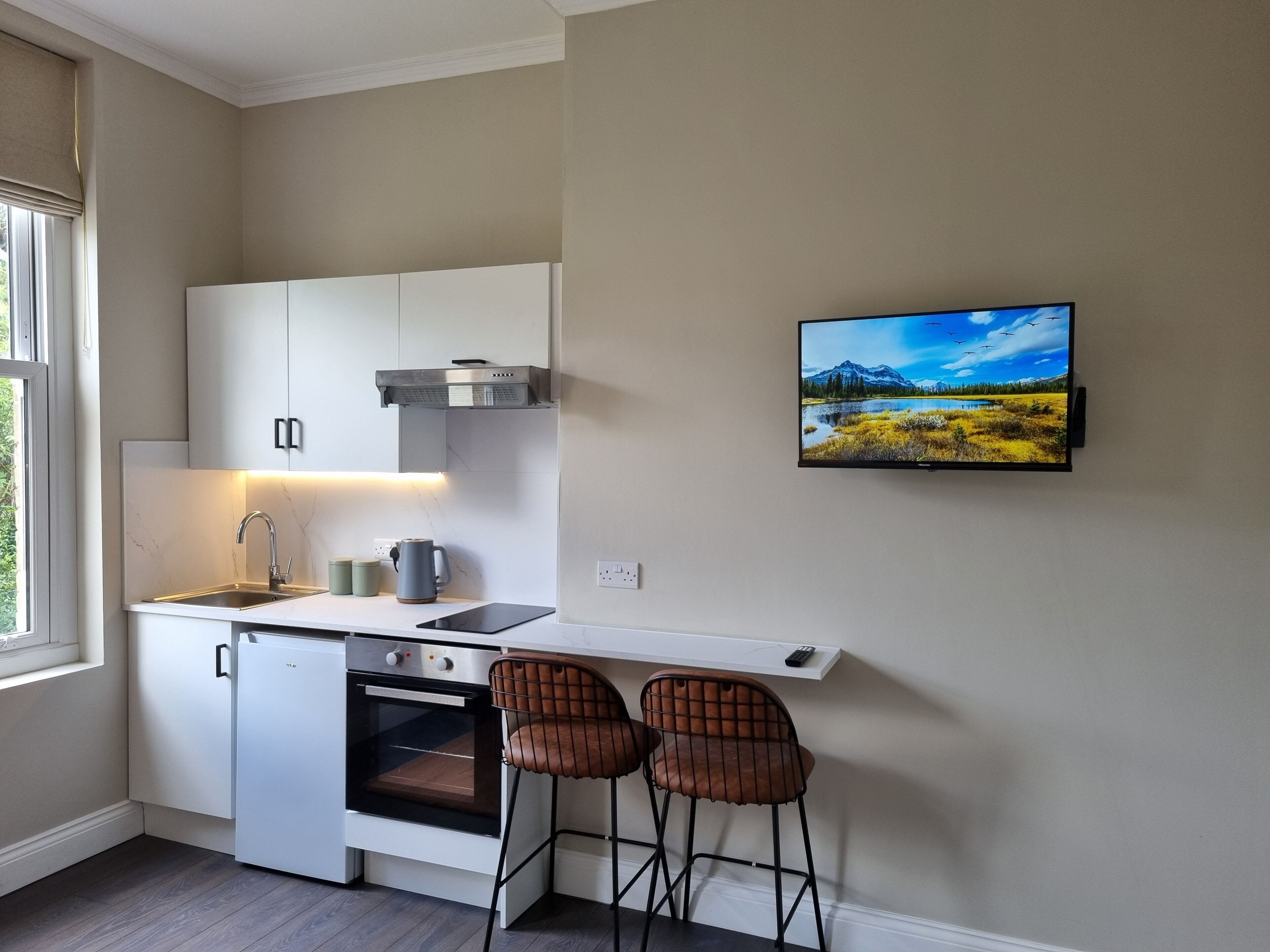 Een compacte keuken met witte kasten, een inbouwoven, een spoelbak en een aanrecht met waterkoker en mokken. Er zijn twee barkrukken en een aan de muur bevestigde tv waarop een schilderachtig landschap te zien is.