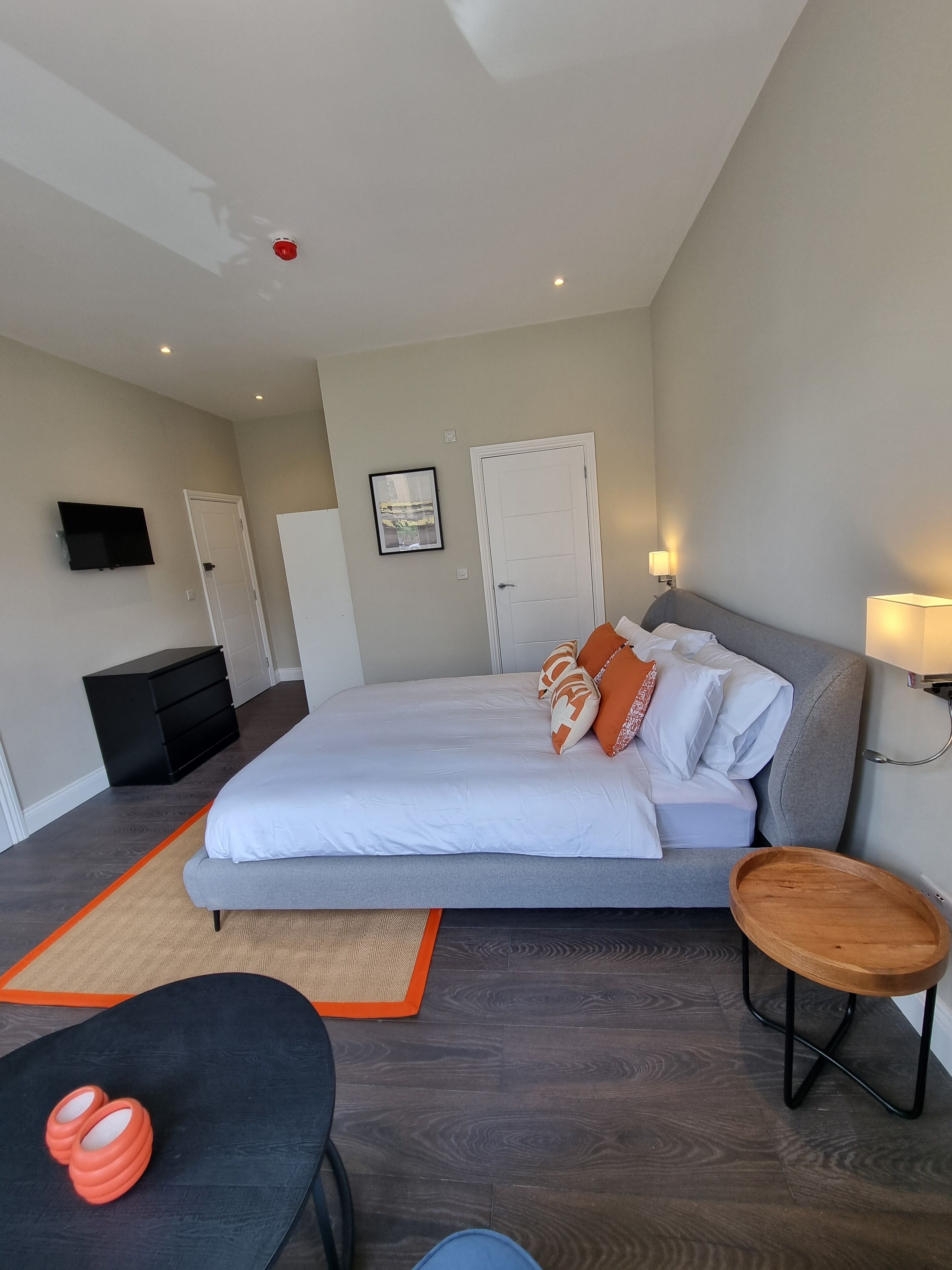 A modern hotel room with a large bed, a desk, a chair, a TV, and a bedside table with a lamp.
