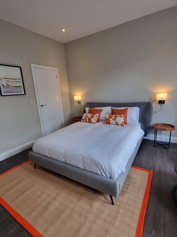 A bedroom with a bed, a nightstand, a wall-mounted lamp, and a door. - The Ivy Apartments (Bournemouth)