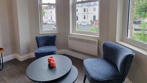 A modern room with two blue armchairs, a round table, and large windows offering a view of the street. - The Ivy Apartments (Bournemouth)