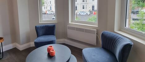 Een moderne kamer met twee blauwe fauteuils, een ronde tafel en grote ramen met uitzicht op de straat.