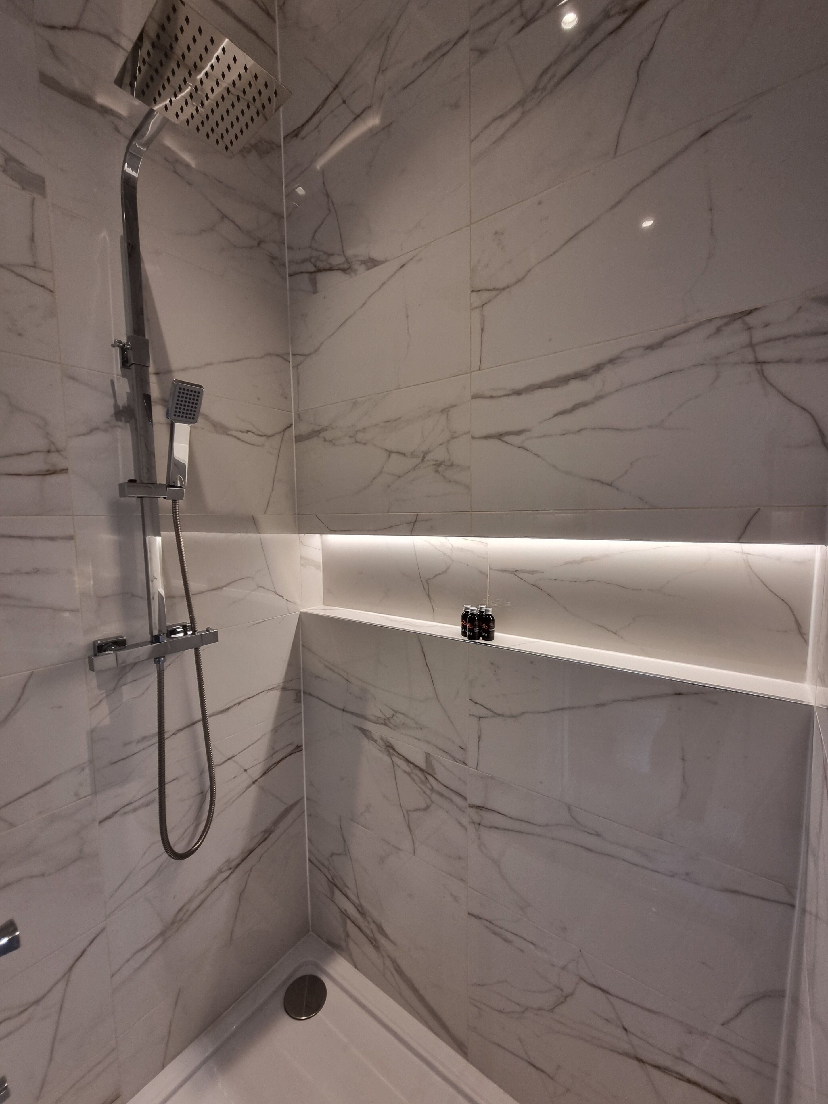 A modern bathroom with a shower, marble walls, and a shelf with bottles.