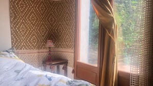 Een slaapkamer met een bed, een nachtkastje, een raam met gordijnen en uitzicht op groen.