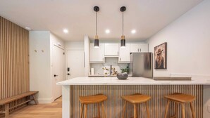 A modern kitchen with a central island, wooden stools, pendant lighting, and a refrigerator.