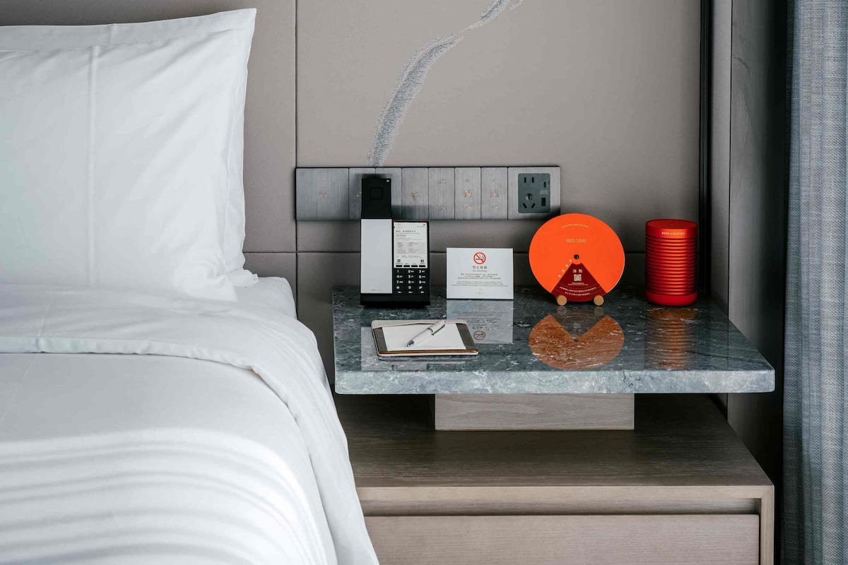 A neatly made bed with white linens, a bedside table with a smartphone, a notepad, a pen, a red cylindrical object, and a wall-mounted light fixture.