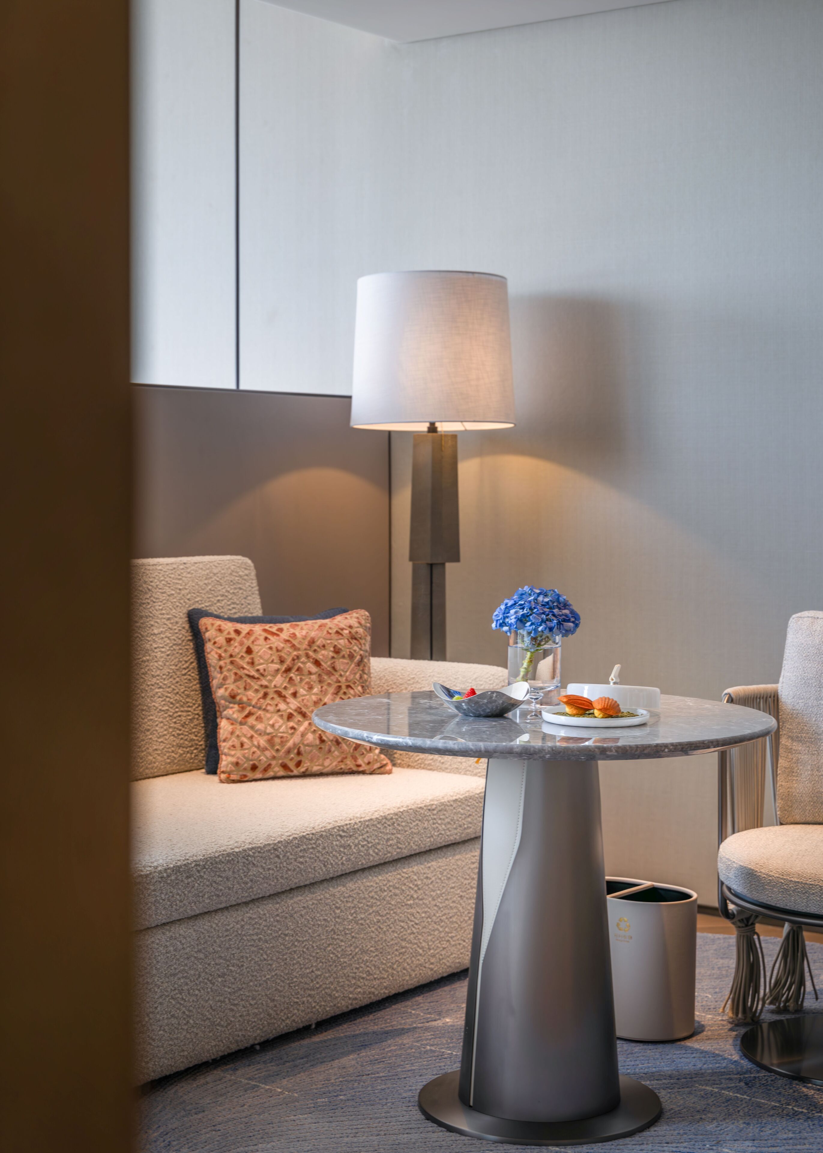 Een moderne hotelkamer met een ronde glazen tafel, een bank, een lamp en een prullenbak.