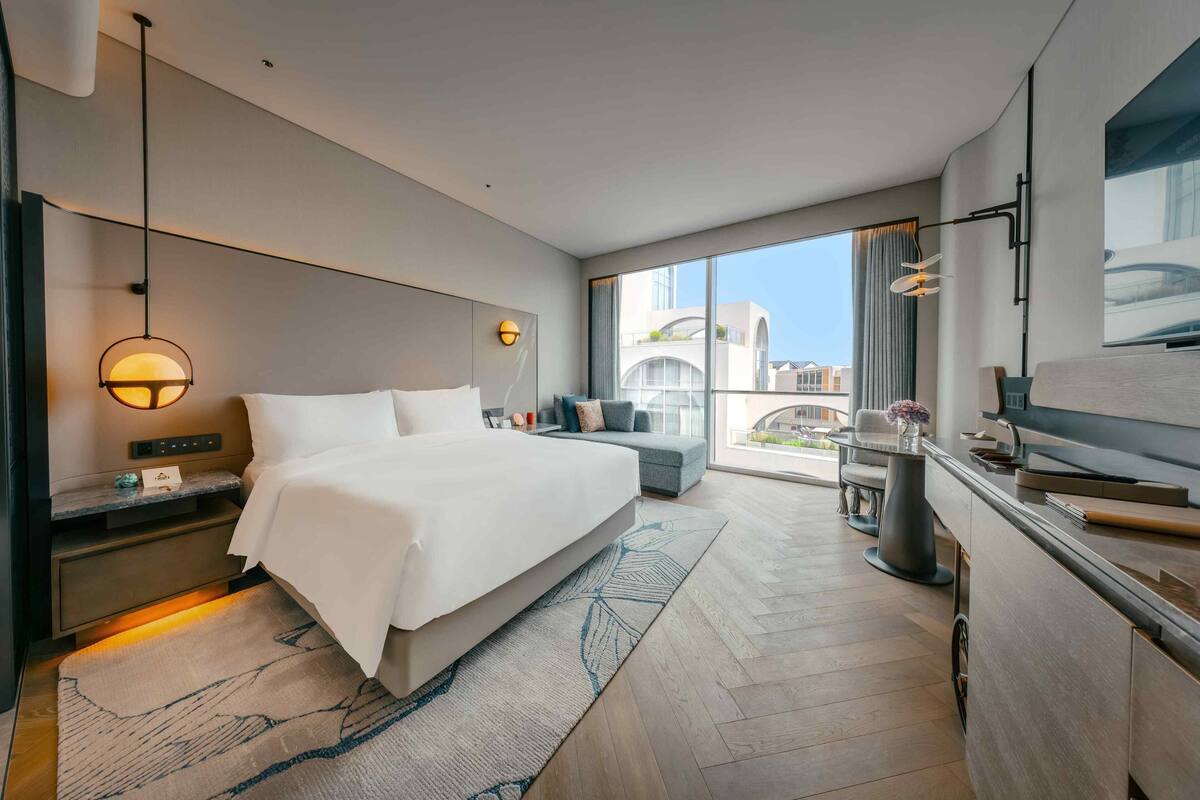 A modern hotel room with a large bed, a desk, a seating area, and a balcony with a view of the city.