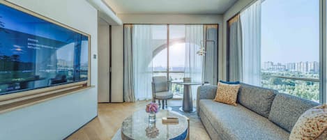 A modern hotel room with a large window, a sofa, a round coffee table, and a TV on the wall.