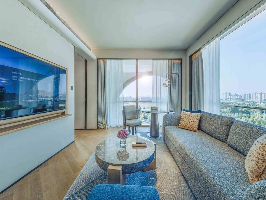 A modern hotel room with a large window, a sofa, a round coffee table, and a TV on the wall.
