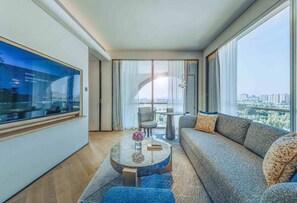 Une chambre d’hôtel moderne avec une grande fenêtre, un canapé, une table basse ronde et un téléviseur fixé au mur.
