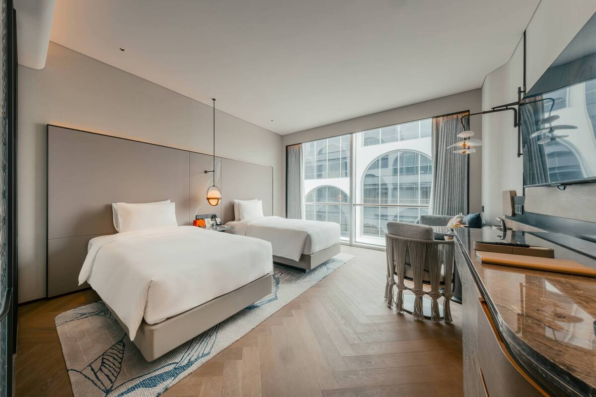 A modern hotel room with two beds, a sitting area with a small table, and a large window offering a view of the city.