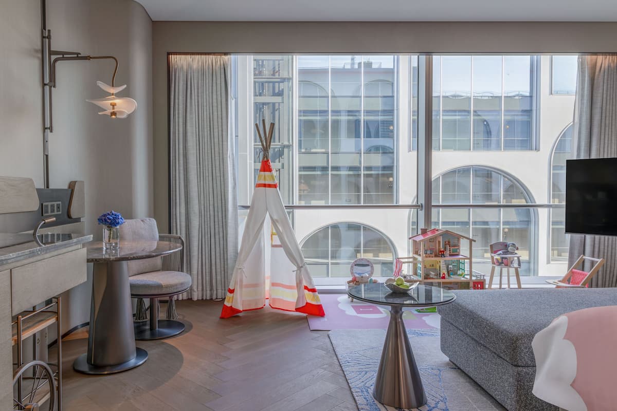 A modern hotel room with a large window, a dining area with a round table, a sofa, a TV, and a play area with toys.