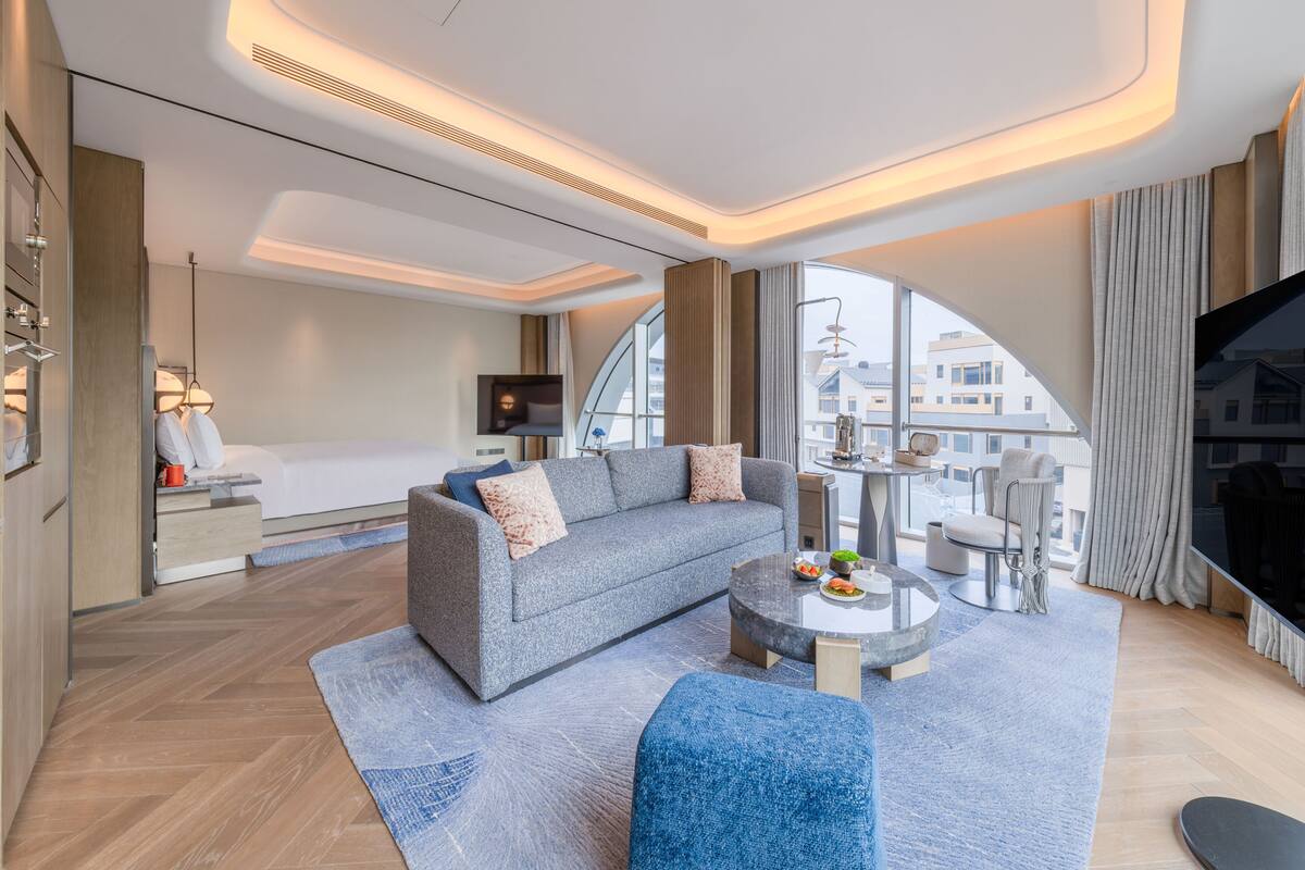 A modern hotel room with a sofa, a round coffee table, a bed, a TV, and a large window offering a view of buildings.
