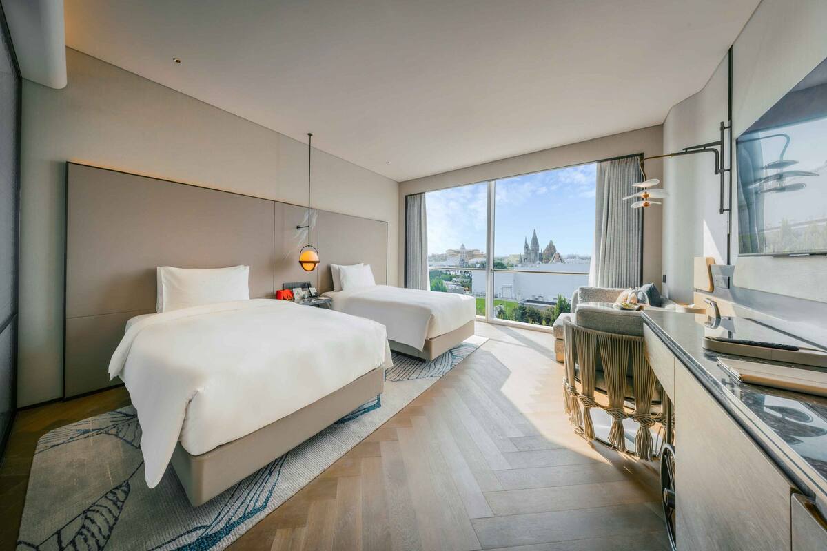 A modern hotel room with two beds, a large window with a city view, a desk with a chair, and a TV.